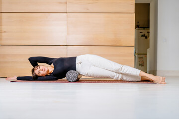 Brunette female yoga instructor in black shirt white trousers costume doing exercise with foam roller for massage in modern cozy home interior. yoga and healthy lifestyle theme concept.