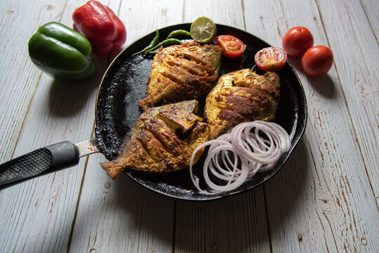Close up of tandoori pomfret fish in a black pan along with ingredients on a background