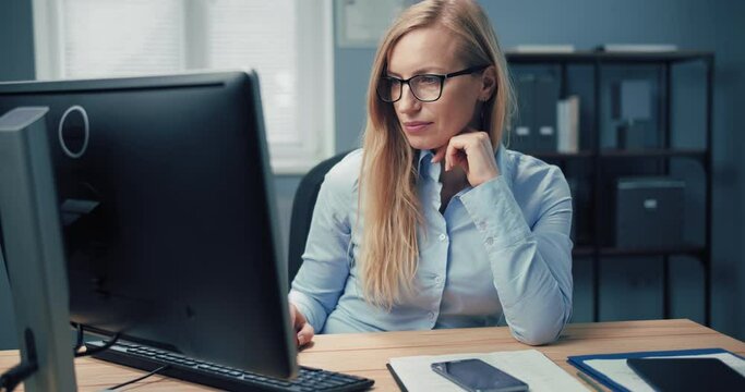 Attractive mature woman in formal wear and eyewear using modern computer while sitting at workplace. Business lady working at bright office with modern device.