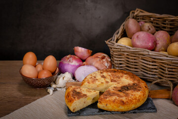 Typical Spanish potato omelette, served at the table with the necessary ingredients for the recipe, potatoes, eggs, onion and garlic