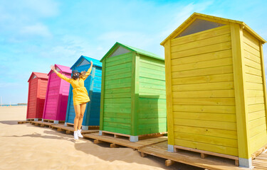 Girl with mask and glasses jumping on the beach with the colored huts next to it. New normal. Concept liberate, joy in the new normal