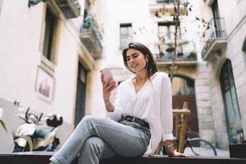 Happy hipster girl in casual clothing resting in courtyard using cellphone gadget for choosing music playlist, carefree Caucasian woman in digital earphones listening positive audio songs from mobile