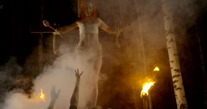 Followers of Satanism from the forest at night bow to the girl crucified on the cross torches burn with fire