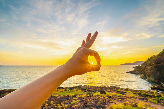The image of a hand posed okay with the sun in the circle. The backdrop is the ocean in the evening, sunset, reflections fell on the calm surface with golden warm light. Feeling refreshed and relaxed.