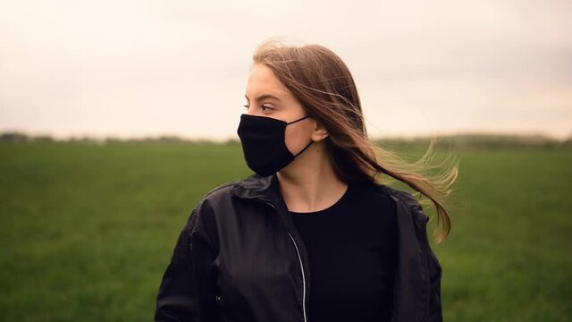A girl in a black jacket and black mask looks at the camera and turns her head to the side against a background of green grass. hair fluttering in the wind. pandemic kovid-19. Coronavirus