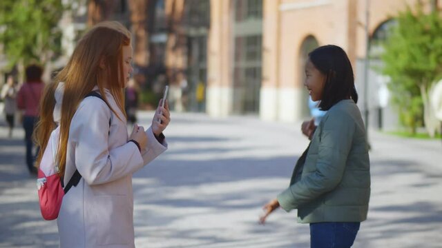 Side view of redhead schoolgirl filming video on phone of asian classmate dancing