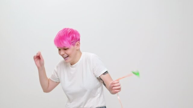 Young beautiful girl with a short haircut and bright hair, painted pink. A homosexual lesbian model poses on a white background in the studio, holds a green balloon in hands.