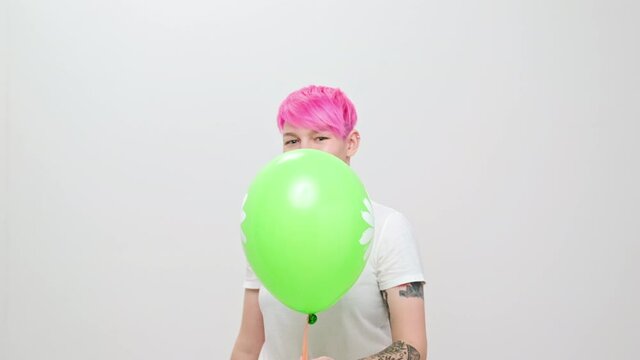 Young beautiful girl with a short haircut and bright hair, painted pink. A homosexual lesbian model poses on a white background in the studio, holds a green balloon in hands.