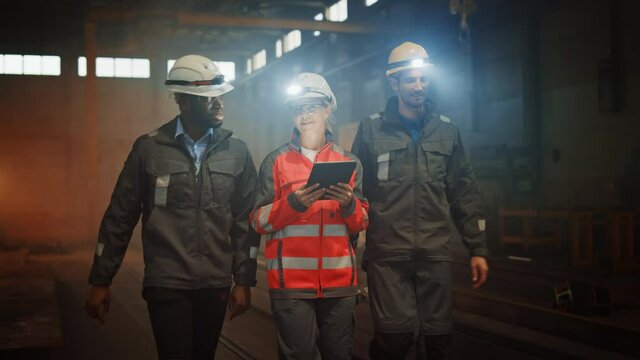 Three Diverse Multicultural Heavy Industry Engineers and Workers in Uniform Walk in Dark Steel Factory Using Flashlights on Their Hard Hats. Female Industrial Contractor is Using a Tablet Computer.