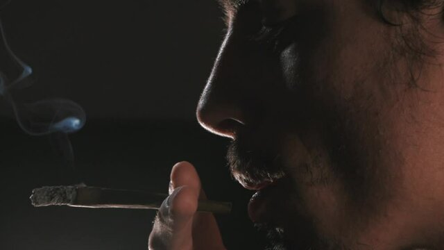Closed frame in slowmotion of a man smoking a CBD blunt with backlight. The end of the shot ends with the smoke from the blunt. ProRes. 4K.
