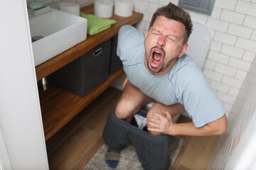 Young man with suffering on his face sits on toilet in bathroom. Indigestion constipation concept.