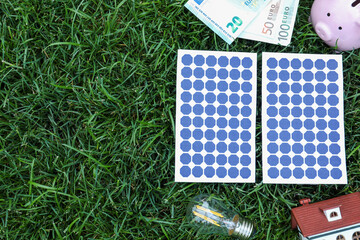 Flat lay composition with solar panels, house model and money on green grass. Space for text