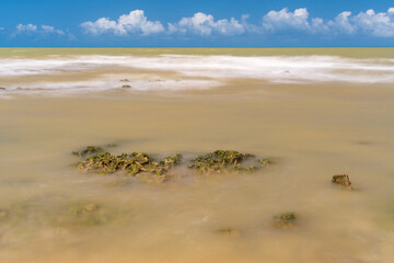 Parts of a reef emerge from a silky sea under a very blue sky full of fluffy and fat clouds.