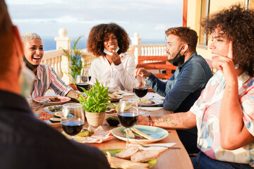 Cheerful multiracial friends enjoy a dinner together outdoor while wearing protective face mask under chin - Coronavirus lifestyle