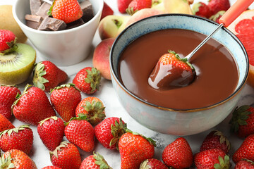 Fondue fork with strawberry in bowl of melted chocolate surrounded by other fruits on table