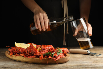 Delicious red boiled crayfishes on wooden table and woman pouring beer into glass on background, closeup