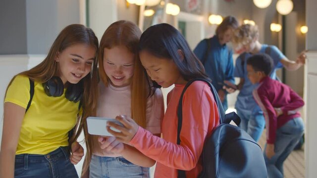 Three multiethnic school girlfriends using smartphone in corridor during break