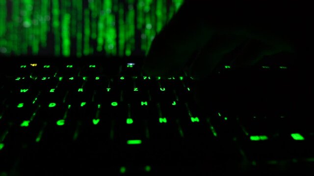 Cybersecurity concept. Hacker entering code on green lit keyboard, hacker code in the background