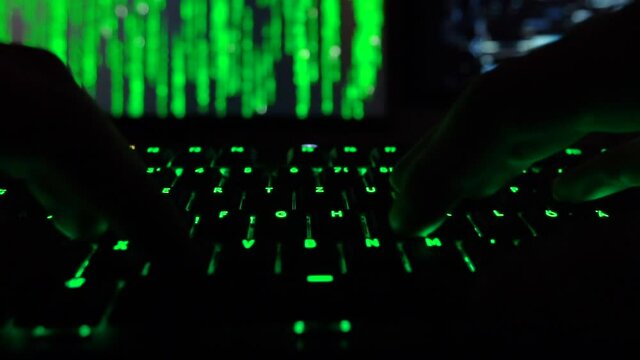 Cybersecurity concept. Hacker in dark room entering code on green lit keyboard, hacker code in the background