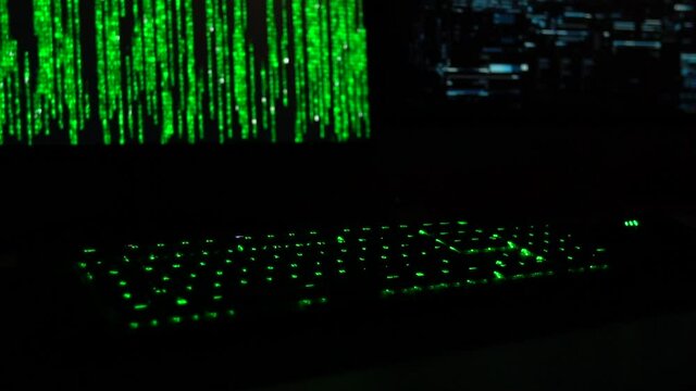 Cybersecurity concept. Slowmotion of green lit keyboard in dark room with hacker code blinking on screen in the background