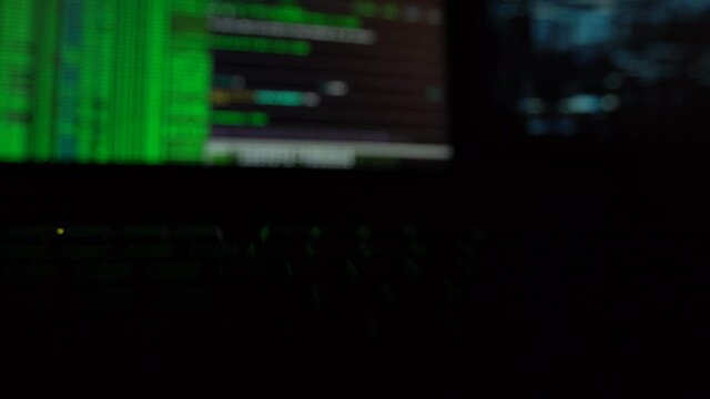 Cybersecurity concept. Closeup of green lit keyboard in dark room with hacker code on screen in the background