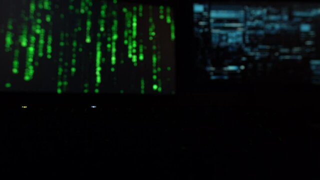 Cybersecurity concept. Closeup of green blinking keyboard in dark room, two screens with hacker code in the background