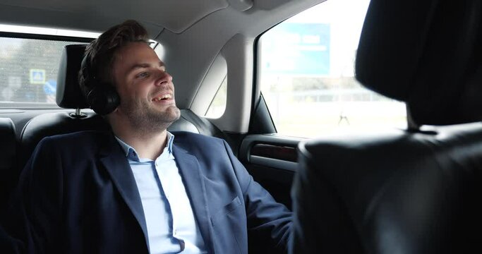 A calm Relaxed Businessman is listening to Music in Business Trip with his own Driver. The man is enjoying the Travel in Business Class Car, wearing Headphones, having a nice Mood. Comfort. Success.