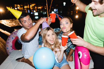 Selective focus of young friends in party caps having fun and holding disposable cups at night