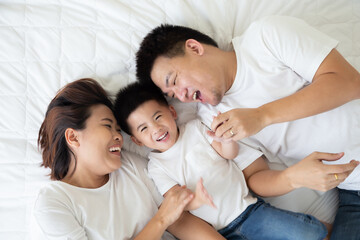 Top view of cute little boy and his asian parents looking at camera and smiling while lying on the white bed at home, Happy Family concept