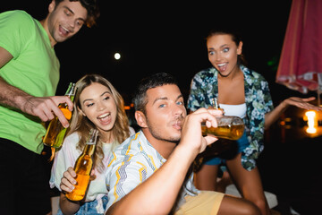 Selective focus of young man drinking beer near excited friends at night