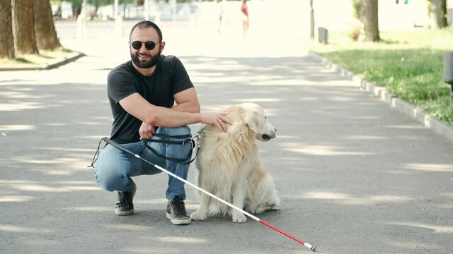 Young blind man is sitting on pavement with a stick and his favorite dog and stroking it. Disabled people life.