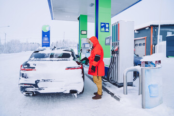 Man in winter red coat holding nozzle with petrol refilling automobile during winter vacation journey, man employee of station providing service to cars