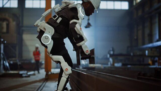Black African American Engineer is Testing a Futuristic Bionic Exoskeleton and Picking Up Metal Objects in a Heavy Steel Industry Factory
