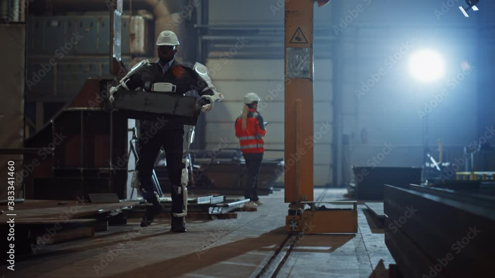 Poster Black African American Engineer is Testing a Futuristic Bionic Exoskeleton and Picking Up Metal Objects in a Heavy Steel Industry Factory. Contractor is Heavy Lifting Steel Parts in a Powered Shell.