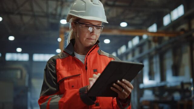 Professional Heavy Industry Engineer/Worker Wearing Safety Uniform and Hard Hat Uses Tablet Computer. Serious Successful Female Industrial Specialist Walking in a Metal Manufacture Warehouse. - Powered by Adobe