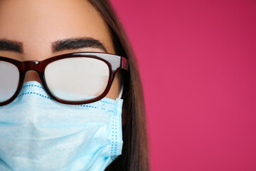 Young woman with foggy glasses caused by wearing disposable mask on pink background, space for...