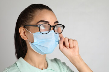 Woman wiping foggy glasses caused by wearing medical mask on light background