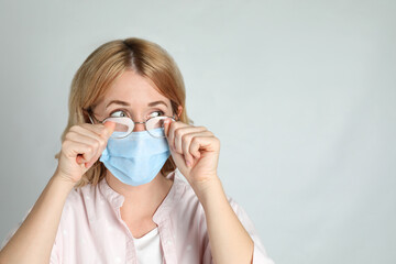 Woman wiping foggy glasses caused by wearing medical mask on light background. Space for text