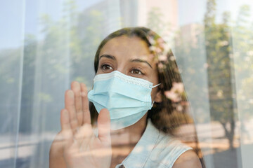 Stressed woman in protective mask looking out of window, view through glass. Self-isolation during COVID-19 pandemic