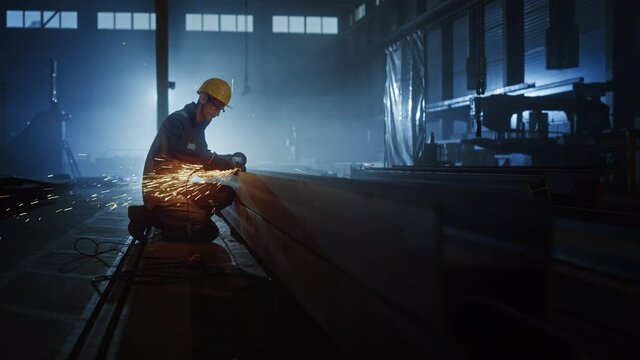 Heavy Industry Engineering Factory Interior with Industrial Worker Using Angle Grinder and Cutting a Metal Tube. Contractor in Safety Uniform and Hard Hat Manufacturing Metal Structures.