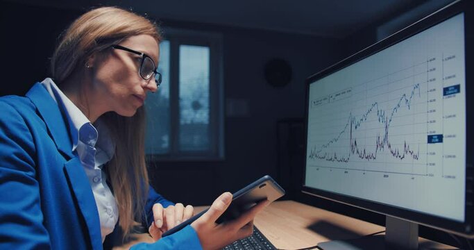 Serious business analyst in eyeglasses and suit preparing financial report on computer. Beautiful lady with blond hair working on project at dark office.
