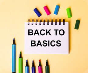 BACK TO BASICS written in a white notebook on a beige background near multi-colored markers
