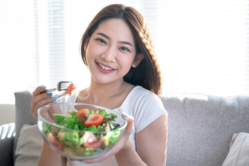 Young happy Asian woman eating healthy salad sitting on the sofa in living room.