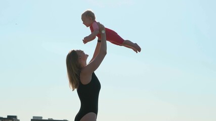 Obraz premium Sexy mother in black closed swimsuit on seashore or ocean throws her daughter up. Child in pink bathing suit. Nice view of happy family on vacation.
