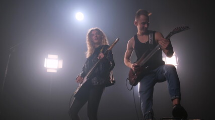 Two rock stars on stage. A lot of light on background. Guy in black T-shirt plays the electric guitar and woman with lush curly hair in leather jacket play too and enjoys music. fog around
