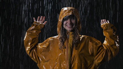 Satisfied woman in yellow raincoat puts on hood raincoat. It is pouring rain, the woman is all wet, she has beautiful wavy wet hair. Lady stands in the street at night.