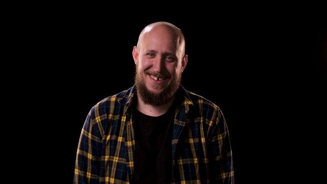 Bald bearded man in checkered shirt looks intently and angrily at the camera on black background. guy smiles and we see that he is missing one front tooth. Funny homeless man on the street at night.