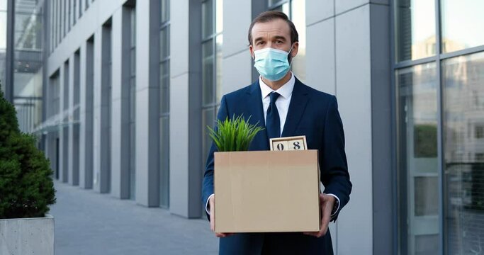 Portrait of retired Caucasian man in medical mask standing outdoor and holding box with personal stuff from office. Senior fired male white-collar worker looking loosing job during pandemic crisis
