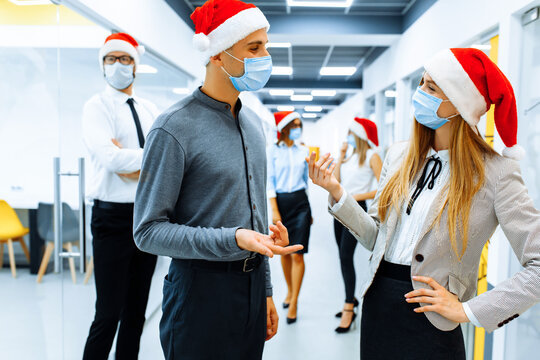 Successful team of business people in medical protective masks on faces and red caps of Santa Claus communicating in the office, Christmas, coronavirus, quarantine