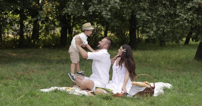 Joyous little boy in a straw hat running towards his smiling parents sitting on the green grass. Happy family concept
